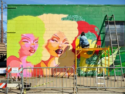 Multidisciplinary artist Vilatose works from scaffolding to complete 'Jardín Cubana,' a striking pop art mural featuring three colorful female figures on a green cinder block wall in Portland's Alberta Arts District. The work-in-progress displays bold neon tones of pink, yellow, and orange against the emerald backdrop, with construction barriers and urban elements framing the creative process under crisp daylight.