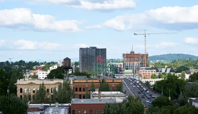 An elevated cityscape view captures Portland's Chinatown district where historic brick buildings meet contemporary high-rise construction. Tower cranes punctuate the skyline as new development rises alongside the ornate classical facade of a heritage building in the foreground. The Burnside Bridge carries steady traffic through the urban corridor, while forested hills provide a verdant backdrop to this evolving neighborhood where preservation and progress intersect.
