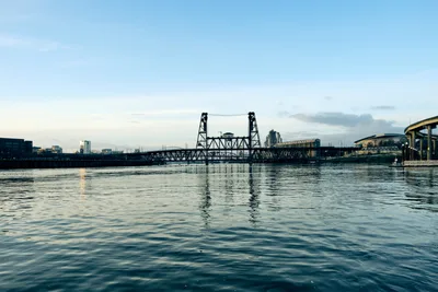 The iconic Steel Bridge spans the Willamette River in downtown Portland, its distinctive lift towers and truss structure creating dramatic silhouettes against a pale afternoon sky. The calm water mirrors the bridge's industrial framework and surrounding cityscape, while soft light catches the rippling surface. Modern buildings and infrastructure frame the scene, capturing the essence of Portland's riverfront where industrial heritage meets contemporary urban development.
