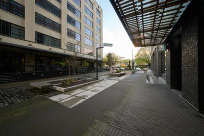 A contemporary pedestrian plaza unfolds between converted industrial buildings in Portland's Pearl District, where geometric brick pavers create flowing pathways beneath a striking steel pergola. The late afternoon light filters through the overhead canopy, casting rhythmic shadows across planters filled with young saplings and modern concrete benches. The juxtaposition of weathered warehouse facades with their grid of steel-framed windows against sleek modern amenities captures the neighborhood's transformation from industrial past to urban living.