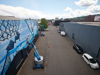 Jeremy Nichols and Devin Finley work from elevated boom lifts to paint an expansive blue and black mural on the side of an industrial building in Southeast Portland's Central Eastside district. The aerial perspective captures the scale of their street art project, with geometric patterns and marine-themed imagery flowing across the concrete wall. Orange safety cones and yellow caution tape mark the work zone on the asphalt below, while scattered vehicles and the urban landscape of warehouses stretch toward forested hills under a partly cloudy sky.