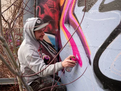 Travis Fields, known as Campographic, applies vivid magenta spray paint to a concrete wall at the Clay Creative Building in Southeast Portland. Clad in a gray hoodie and cap, the muralist works amid bare winter branches, adding electric pink highlights to an existing composition of flowing forms and geometric elements. The industrial setting and natural light create an authentic documentary moment of urban art creation in Oregon's creative district.