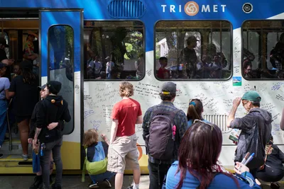 Community members gather around a retiring TriMet Type 1 MAX train in Portland's Lloyd District, transforming its white exterior into a canvas of handwritten messages and memories. Passengers peer through windows while dozens of people of all ages write personal tributes on the train's surface, creating an impromptu memorial to decades of public transit service. The scene captures both celebration and nostalgia as Portland bids farewell to its original light rail fleet on a bright spring afternoon.