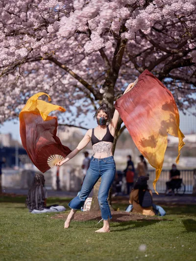 A masked performer dances gracefully with flowing orange and red silk scarves beneath the peak bloom of cherry blossoms at Tom McCall Waterfront Park. The dancer, wearing a black top and jeans, moves barefoot on the grass while park visitors relax in the background under the canopy of pink sakura petals. Golden hour light filters through the blossoms, creating a dreamy atmosphere that captures the ephemeral beauty of Portland's spring season.