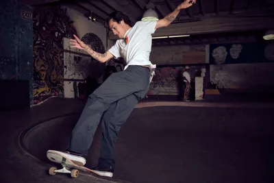 A tattooed skateboarder carves through the concrete bowl at Commonwealth Skateboarding in Portland's Buckman neighborhood, his body extended in fluid motion against graffiti-covered walls. The underground skate park's moody lighting creates dramatic shadows as he navigates the curved transition, embodying the raw energy and artistic expression of Portland's skating culture. His crisp white shirt and relaxed stance demonstrate effortless control within the gritty, art-adorned environment.