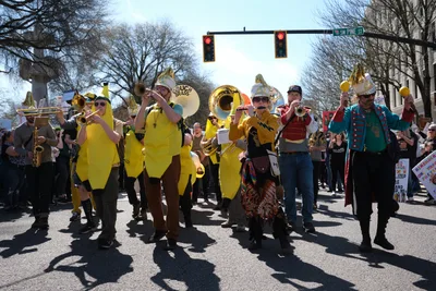 The Unpresidented Brass Band commands Southwest Pine Street in Portland's Old Town district, their golden instruments gleaming against a backdrop of bare winter branches and traffic signals glowing red. Musicians in vibrant yellow costumes and eclectic attire march purposefully through the intersection, their brass section creating a defiant soundtrack for the March 2026 No Kings Protest. Sharp shadows stretch across the asphalt while onlookers gather along the sidewalks, witnessing this spirited demonstration of musical activism in the heart of downtown Portland.