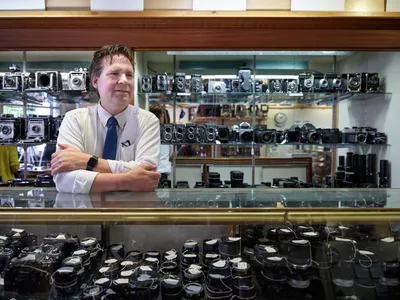 Mike Knight stands confidently behind the glass display case at Blue Moon Camera & Machine, his arms crossed in a gesture of quiet authority over his domain of vintage cameras and lenses. The meticulous arrangement of countless film cameras on illuminated shelves creates a cathedral-like atmosphere of photographic history, while warm overhead lighting bathes the scene in golden tones that reflect off the polished glass surfaces. Knight's professional attire—crisp white shirt and navy tie—contrasts beautifully with the mechanical artistry surrounding him, embodying the intersection of traditional craftsmanship and modern retail expertise in Portland's Saint Johns neighborhood.