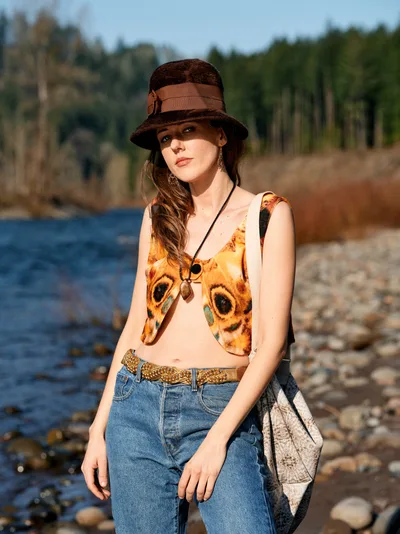 A young woman in vintage-inspired attire poses along the rocky riverbed of the Floodplain Trail in Multnomah County, Oregon. She wears a distinctive graphic top featuring bold owl eyes, paired with high-waisted denim and a chocolate brown fedora hat. The warm golden hour light illuminates her contemplative expression as she stands against the backdrop of the Columbia River's gentle waters and forested hills.