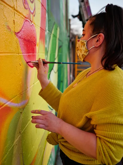 Multidisciplinary artist Vilatose applies precise brushstrokes to her Jardín Cubana mural depicting three female figures along Northeast Alberta Street in Portland's vibrant arts district. Wearing a coordinating mustard sweater and floral face mask, she works with focused concentration against the kaleidoscopic backdrop of existing street art. The warm afternoon light illuminates both her careful technique and the textured concrete canvas that defines this creative corridor.