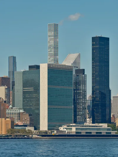 The iconic Manhattan skyline rises majestically from the East River, captured from Brooklyn's Greenpoint neighborhood on a crystalline afternoon. The United Nations Headquarters' distinctive rectangular form anchors the composition alongside gleaming residential towers, their glass facades catching the clear light against an azure sky. Gentle ripples disturb the river's surface in the foreground, while wispy clouds drift across the expansive urban panorama. The interplay of modernist architecture and classical skyscraper forms creates a compelling study of New York's evolving vertical landscape.
