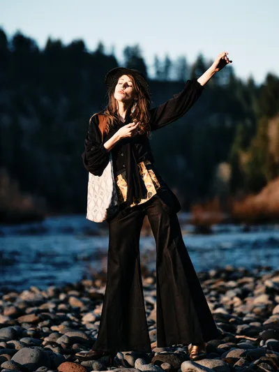 A young woman in flowing black garments and a wide-brimmed hat strikes an expressive pose on the rocky riverbed of the Columbia River along Oregon's Floodplain Trail. The golden hour light bathes her figure as she extends one arm skyward while her other hand rests on her chest, creating a moment of theatrical grace against the silhouetted forest backdrop. Her layered outfit includes flowing wide-leg pants and a loose jacket over a white top, with warm sunlight catching the fabric's movement. The composition places her among smooth river stones with the darkened treeline of Multnomah County creating dramatic contrast in the background.