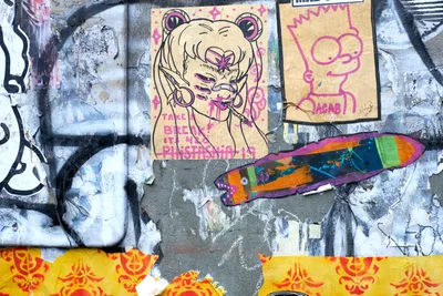 A vibrant wheat paste art installation adorns a weathered concrete wall in Portland's Central Eastside district. The composition features a whimsical anime-inspired character with bear ears surrounded by pink decorative elements, alongside colorful fish motifs and simple line drawings. Layers of torn and overlapping paper create a rich textural collage against the industrial urban backdrop, with yellow and red stenciled elements anchoring the bottom of the frame.