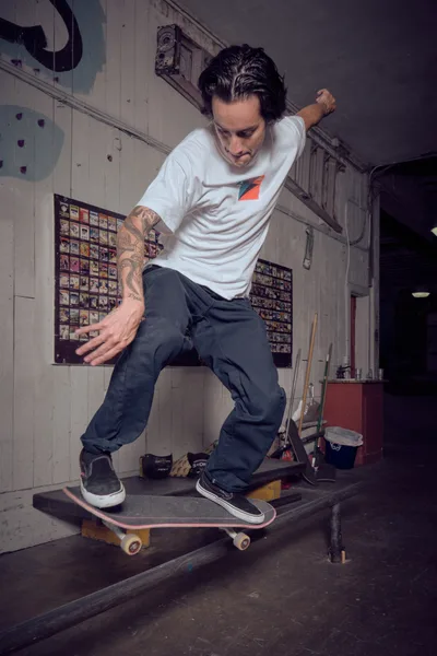 A tattooed skateboarder in a white tee and dark jeans demonstrates precise board control inside Commonwealth Skateboarding in Portland's Buckman neighborhood. The intimate shop setting reveals walls lined with skateboard decks and weathered industrial fixtures, while warm ambient lighting captures the focused concentration and fluid motion of street skating culture. The rider's stance suggests years of practiced balance, frozen mid-maneuver against the authentic backdrop of Portland's skateboarding community.