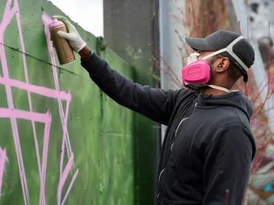 A focused muralist in protective respirator and cap applies vibrant green and pink spray paint to a concrete wall in the parking lot of Clay Creative in Portland's Hosford-Abernethy neighborhood. The artist, wearing a dark hoodie and white gloves, works with methodical precision against the backdrop of existing graffiti art. The industrial setting captures the raw creative energy of Southeast Portland's evolving street art scene.