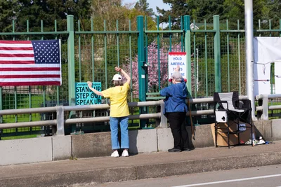 Two activists position handmade protest signs along the concrete barrier of the North Skidmore Street overpass in Portland, their messages visible to Interstate 5 traffic below. One demonstrator in a bright yellow shirt raises their arm toward passing vehicles, while their companion in navy blue secures additional signage. The scene unfolds against spring foliage with an American flag prominently displayed alongside calls for civic engagement, creating a tableau of grassroots political expression in urban Oregon.