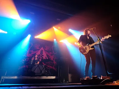Emma Richardson commands the foreground on bass guitar while Julian Dorio anchors the rhythm from behind his drum kit during Band of Skulls' 10 Year Anniversary Tour at Webster Hall. Theatrical stage lighting cuts through the darkness in brilliant beams of blue, amber, and white, creating a dramatic atmosphere that illuminates the duo against their backdrop. The intimate Manhattan venue's exposed ceiling beams frame the performance as colored spotlights cascade across the musicians, capturing the raw energy of this milestone celebration.