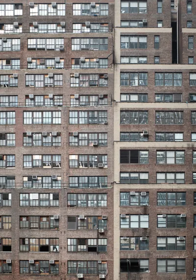 A dense pattern of weathered brick apartment building facades creates an urban tapestry in Midtown Manhattan. The repetitive grid of windows, punctuated by air conditioning units and varying states of wear, reveals the intimate details of city living through glimpses of curtains, blinds, and personal belongings. The warm brown and red brick tones contrast with the cool reflective glass, while the geometric composition emphasizes the vertical density of New York residential architecture.