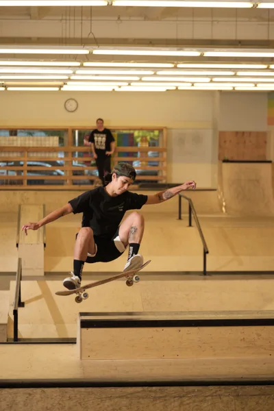 Professional skateboarder Christiana Means executes a weightless maneuver above the wooden transitions of Stronger Skatepark in Milwaukie, Oregon. The harsh fluorescent lighting casts dramatic shadows across the blonde plywood surfaces while another skater observes from the background. Her tattooed form is frozen mid-air, skateboard locked beneath her feet in a moment of athletic grace against the utilitarian architecture of the indoor facility.