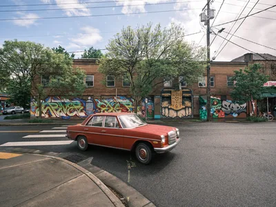 A crimson vintage Mercedes-Benz sedan glides through the intersection at Southeast Division Street in Portland's Richmond neighborhood, its classic lines contrasting against a vibrant wall of street art adorning the former Oregon Theater. The weathered brick building serves as an urban canvas, its colorful graffiti murals creating a dynamic backdrop under the dappled afternoon light filtering through mature street trees. Power lines cross overhead like geometric sketches against the soft blue sky, while the vintage automobile becomes a moving piece of the neighborhood's evolving artistic identity.