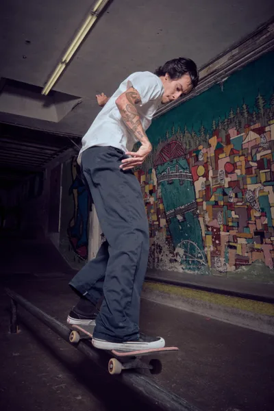 A tattooed skateboarder in white t-shirt and dark pants navigates a weathered skateboard through an atmospheric underground tunnel at Commonwealth Skateboarding in Portland's Buckman neighborhood. The industrial space features exposed fluorescent lighting and a vibrant mural depicting geometric patterns and urban landscapes across the wall. The moody lighting creates dramatic shadows while highlighting the rider's concentrated expression and fluid movement through this gritty subterranean environment.