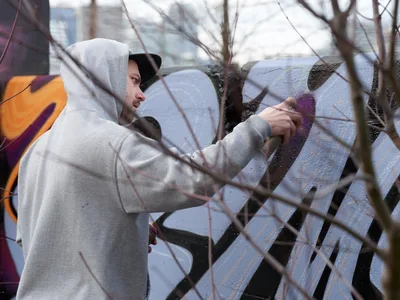 Travis Fields, known as Campographic, works intently on a vibrant mural at the Clay Creative Building in southeast Portland's Hosford-Abernethy neighborhood. Viewed through bare winter branches, the hooded artist applies purple paint to the concrete wall while afternoon light filters through the urban landscape. The scene captures the intersection of creative expression and community space, with colorful graffiti art visible in the background.