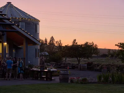 Golden hour bathes a converted grain silo home at Abbey Road Farm in Carlton, Oregon, as a small group mingles on the patio beneath string lights. The corrugated metal structure features a distinctive conical roof crowned with fairy lights, while the salmon-pink sunset sky creates a warm backdrop against the Willamette Valley landscape. Wine barrels and rustic landscaping complete this intimate agricultural gathering space.