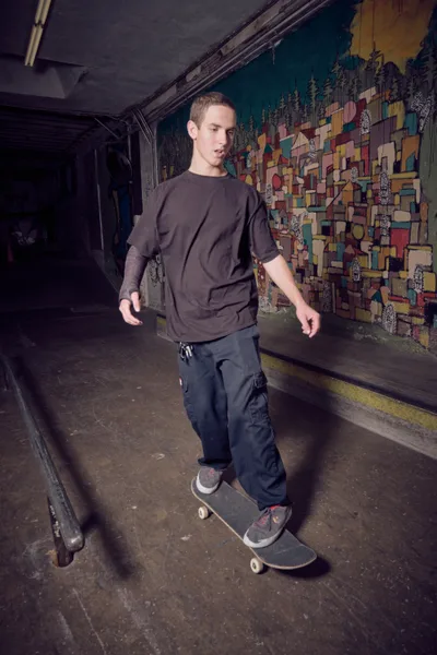A young skater glides through the dimly lit passage at Commonwealth Skateboarding in Portland's Buckman neighborhood. His dark streetwear silhouette moves against a vibrant geometric mural that transforms the tunnel walls into an explosion of color and pattern. The industrial concrete setting creates a stark contrast with the artistic backdrop, while fluorescent lighting casts dramatic shadows that emphasize the raw, authentic atmosphere of Portland's underground skate scene.