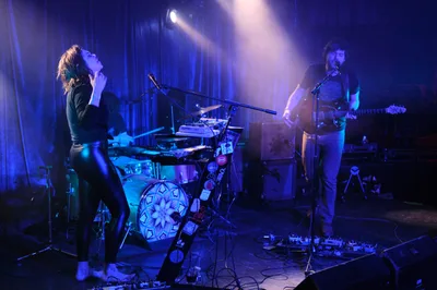 Experimental band Sun Blood Stories delivers an electrifying performance at Rx Fest in the intimate confines of Dante's nightclub in downtown Portland. Stage lights pierce through swirling atmospheric haze, casting the Boise-founded duo in dramatic purple and blue hues as the vocalist throws back their head in passionate delivery while the guitarist anchors their mesmerizing sound. The low-ceiling venue creates an intensely personal connection between performers and audience, with cables snaking across the worn floor and the drummer's ornate kick drum design catching the moody lighting.