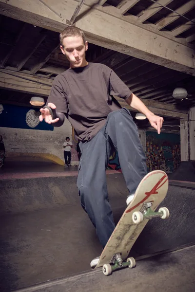 A young skater demonstrates precise balance and control while riding through the weathered concrete bowls of Commonwealth Skateboarding in Portland's Buckman neighborhood. The industrial atmosphere is captured through exposed wooden ceiling beams and worn concrete surfaces, while warm artificial lighting creates dramatic shadows that emphasize the fluid motion of the skateboard beneath his feet. The skater's concentrated expression and relaxed posture suggest years of practice in this underground sanctuary.