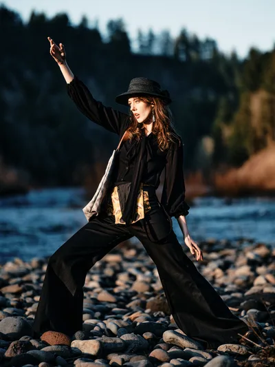 A young woman in flowing black attire strikes a dramatic pose on the rocky riverbed of Oregon's Columbia River. Her dark hat and billowing garments catch the golden hour light as she extends one arm skyward in theatrical expression. The forest-lined Floodplain Trail creates a moody backdrop, with weathered river stones beneath her feet and the gentle current beyond.