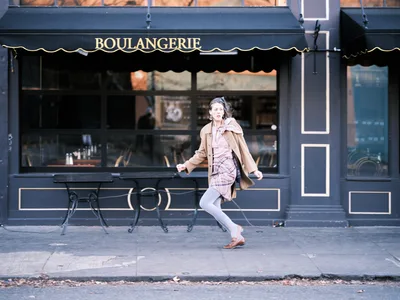 A woman in motion strides past a classic French bakery storefront on Northeast Alberta Street in Portland's Alberta district. Her windswept hair and flowing camel coat capture the energy of city life as she hurries along the sidewalk, while the dark navy boulangerie facade with its elegant gold lettering and wrought iron outdoor seating creates a timeless European atmosphere. The morning light catches her pink plaid dress and creates a dynamic street photography moment that embodies urban Portland's blend of old-world charm and contemporary pace.
