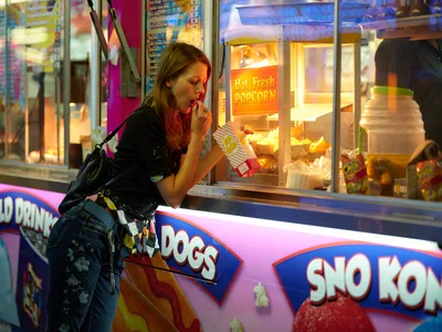 A young woman with long brown hair leans toward a glowing popcorn stand at the Clark County Fair in Vancouver, Washington, her hand thoughtfully raised to her mouth as she contemplates the tempting snack. The warm amber light from the concession stand illuminates her profile against the vibrant carnival atmosphere, while colorful signage for hot dogs and sno-kones creates a nostalgic backdrop. The scene captures the quintessential fairground moment of indulgent decision-making amid the sensory overload of carnival treats and neon lights.