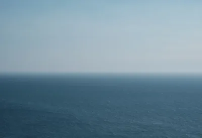 A minimalist seascape captures the infinite meeting of steel-blue Pacific waters and pale gray sky along the Oregon coastline. The horizon line divides the frame with mathematical precision, creating a meditative study in monochromatic blues and grays. Subtle texture in the ocean's surface hints at gentle swells, while the overcast sky maintains an ethereal, weightless quality typical of the Pacific Northwest's marine climate.