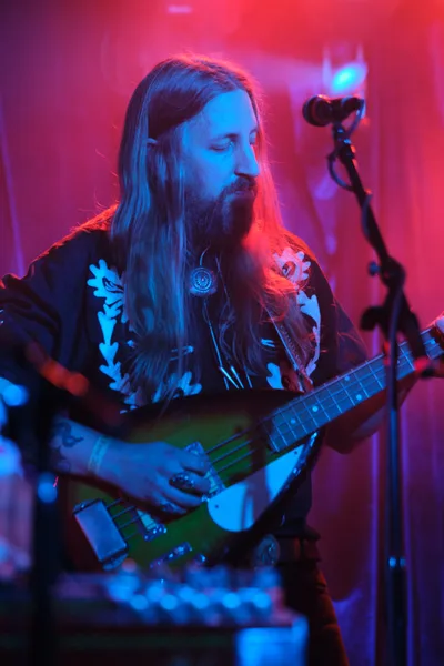 Tobias Berblinger of Roselit Bone commands the intimate stage at Dante's in Portland, his flowing hair catching the electric interplay of magenta and cyan stage lighting. The bassist's weathered hands work the fretboard of his instrument while atmospheric smoke and colored gels transform the legendary venue into a dreamscape of saturated hues. His ornate black shirt adorned with floral motifs speaks to the band's theatrical aesthetic, while the concentrated expression on his bearded face reveals an artist lost in the sonic landscape of RX Fest 2019.