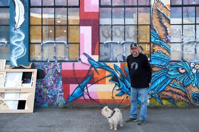 A bearded middle-aged man in black clothing and baseball cap stands confidently beside his small white terrier against a vibrant street mural in Portland's Central Eastside. The expansive graffiti artwork features dramatic blue feathers and mythical creatures painted across boarded windows, creating a striking urban canvas. Natural daylight illuminates the scene, highlighting the contrast between the artist's casual presence and the bold, colorful street art that transforms this industrial neighborhood into an outdoor gallery.