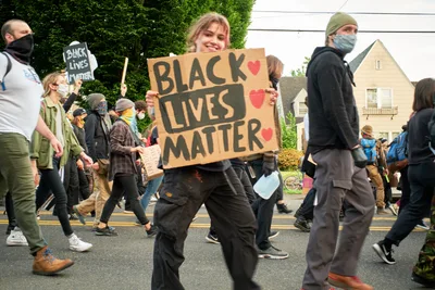 Demonstrators stride purposefully down East Burnside Street in Portland's Laurelhurst neighborhood during a Black Lives Matter protest following George Floyd's murder. At the center, a young woman with blonde hair carries a handmade cardboard sign reading 'BLACK LIVES MATTER' adorned with pink hearts, her expression radiating determination and hope. The crowd moves as one through the tree-lined residential street, masked faces reflecting the pandemic era's intersection with social justice activism. Suburban homes with peaked roofs frame the procession, creating an intimate contrast to the movement's national scale.