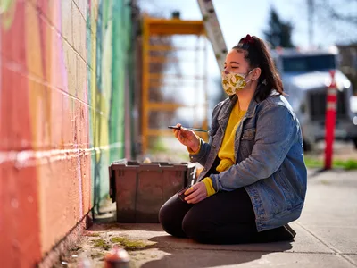 Multidisciplinary artist Vilatose sits cross-legged on the sidewalk, painting brush in hand, working on Jardín Cubana, a vibrant mural depicting three female figures along Alberta Street in Portland's historic arts district. Wearing a yellow patterned face mask and denim jacket over a mustard sweater, she pauses in her creative process against the colorful backdrop of pink, coral, and green paint strokes. Golden afternoon light bathes the scene, highlighting the weathered concrete and moss-touched pavement of this iconic Northeast Portland creative corridor.
