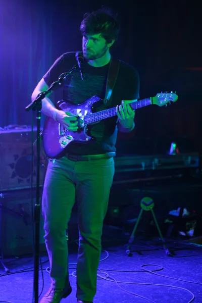 A bearded musician in a black t-shirt commands the intimate stage at Dante's in downtown Portland, his electric guitar caught mid-chord under saturated purple and green stage lighting. The atmospheric wash of color transforms the underground venue into a moody sanctuary where Sun Blood Stories weaves their sonic narrative. Cable-strewn floors and shadowed equipment create texture in the foreground while the performer's concentrated expression speaks to the focused intensity of this experimental band's mesmerizing live sound. Originally from Boise, Idaho and now Portland-based, the group channels their operatic approach to music through every chord.