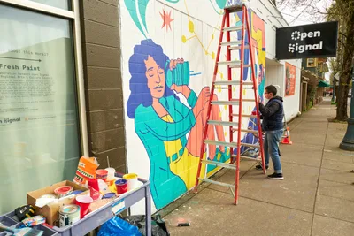 Artist Anke Gladnick works on a vibrant community mural titled "Let's Talk" on the exterior wall of Open Signal in Portland's Eliot neighborhood. The colorful artwork depicts a figure with blue hair holding what appears to be a vintage camera or recording device, rendered in bright teals, yellows, and coral tones. A red stepladder stands against the wall as the artist adds finishing touches, while paint cans and supplies are organized on a nearby table, capturing the collaborative spirit of public art creation.