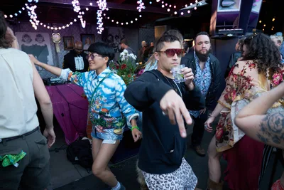 Revelers lose themselves in the rhythm at Your Sunday Best day party, bathed in the moody purple glow of string lights cascading from White Owl Social Club's ceiling. A woman in a vibrant tropical print shirt dances with abandon while a man in sunglasses and black clothing extends his hand toward the camera, creating an intimate connection with the viewer. The crowded dance floor pulses with eclectic fashion choices and uninhibited movement, capturing the essence of Portland's underground party culture in this dimly lit, art-adorned venue.
