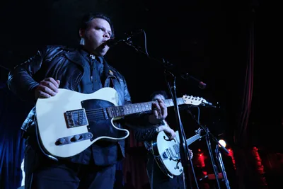 Victor Franco of Roselit Bone commands the shadowed stage at Dante's, his white Telecaster gleaming against the venue's inky atmosphere. Bathed in moody blue stage lighting with crimson accents bleeding from the background, Franco leans into the microphone with focused intensity while his bandmate's hollow-body guitar catches fragments of light in the periphery. The intimate Portland club setting creates a cocoon of sound and shadow, where the performer's leather jacket and concentrated expression speak to the raw authenticity of the underground music scene.