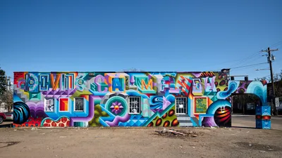 A vibrant geometric mural explodes across the entire facade of a single-story building in Portland's Alberta district, creating a mesmerizing tapestry of interlocking shapes and saturated colors. The street art features bold typography spelling "ALIVE" alongside abstract forms in electric blues, magentas, oranges, and greens that seem to pulse against the crystalline winter sky. Power lines slice horizontally through the composition while the building's original windows peek through the psychedelic geometry like portals.