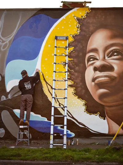 Street artist Travis Fields, working under the name campographic, applies vibrant paint to a towering mural of poet Amanda Gorman on a brick wall in Portland's Alberta district. The artwork depicts Gorman with an upward gaze against a brilliant yellow sun and flowing blue waves, while Fields balances on a stepladder, his dark hoodie contrasting against the warm tones of his creation. An aluminum extension ladder stretches upward along the wall, with paint cans scattered on the grass below, capturing the active process of public art creation in this Northeast Portland neighborhood.
