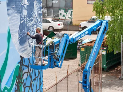 Jeremy Nichols and Devin Finley collaborate on a monumental street art installation in Southeast Portland's Central Eastside district, their blue Genie lift extending toward a towering wall mural featuring flowing abstract forms in blues and greens. The industrial urban backdrop includes chain-link fencing, parked vehicles, and verdant spring foliage, capturing the intersection of artistic creation and city infrastructure. Golden afternoon light illuminates their focused work, documenting the Portland Street Art Alliance's commitment to transforming public spaces through large-scale community art.