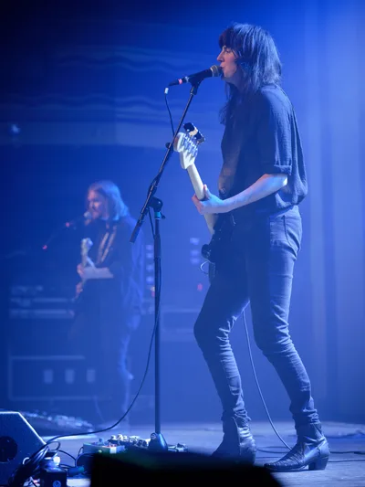 Emma Richardson of Band of Skulls performs under saturated blue stage lighting at Webster Hall in Manhattan, her silhouette defined against the moody atmospheric glow. The intimate venue setting captures the raw energy of the anniversary tour, with Richardson positioned at center stage while bandmate Russell Marsden remains partially visible in the background shadows. Her focused expression and commanding stage presence anchor this moment of live musical performance in the iconic East Village venue.