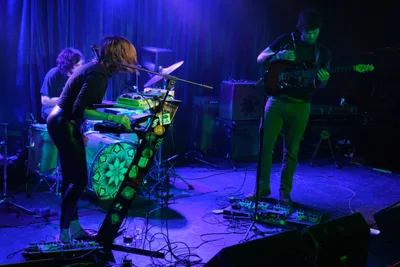 Sun Blood Stories delivers an intimate performance bathed in electric blue and green stage lighting at Dante's in downtown Portland. The keyboardist leans into their craft at center stage, flanked by intricate mandala artwork on their instrument, while the guitarist commands the right side of the frame. Cables snake across the wooden floor of this legendary venue, creating geometric patterns that mirror the psychedelic atmosphere of the underground music scene.