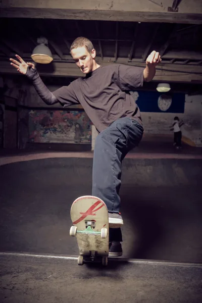 A young skater demonstrates perfect balance while riding a distinctive red lightning bolt-decorated skateboard through the gritty interior of Commonwealth Skateboarding in Portland's Buckman neighborhood. His arms extend gracefully for stability as he navigates the worn concrete floors beneath exposed ceiling beams and industrial lighting. The atmospheric shot captures the raw authenticity of skate culture within this iconic Southeast Portland skate shop, where weathered surfaces and graffitied walls create a backdrop steeped in street credibility.
