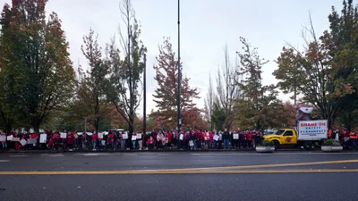 A sea of red-clad nurses and supporters mass along Northeast Multnomah Street outside Kaiser Permanente Tower in Portland's Lloyd District, their protest signs creating a rhythmic pattern against autumn's burnished foliage. The overcast Pacific Northwest sky casts diffused light across the demonstration, while a yellow union truck bearing "SHAME ON KAISER PERMANENTE" anchors the right side of the frame. The striking healthcare workers stretch in an unbroken line along the sidewalk, their collective presence transforming the corporate streetscape into a theater of labor solidarity.