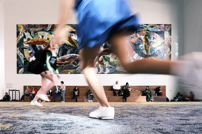 Blurred figures in motion streak across the foreground as visitors hurry through the spacious foyer of Portland's Oregon Convention Center. Behind them, spectators rest on terraced concrete steps beneath a vibrant, large-scale mural featuring serpentine forms and bold street art aesthetics. The juxtaposition of kinetic energy and static contemplation creates a dynamic urban tableau, with warm lighting illuminating the contemporary architectural space.