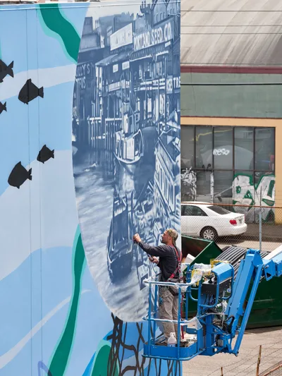 Jeremy Nichols and Devin Finley work from a blue scissor lift to paint an expansive mural on a warehouse wall in Southeast Portland's Central Eastside district. The marine-themed artwork features silhouetted fish swimming through aqua waters alongside a detailed blue-toned maritime scene. Bright afternoon sunlight illuminates their progress on this large-scale public art installation, part of the Portland Street Art Alliance's ongoing urban beautification efforts.